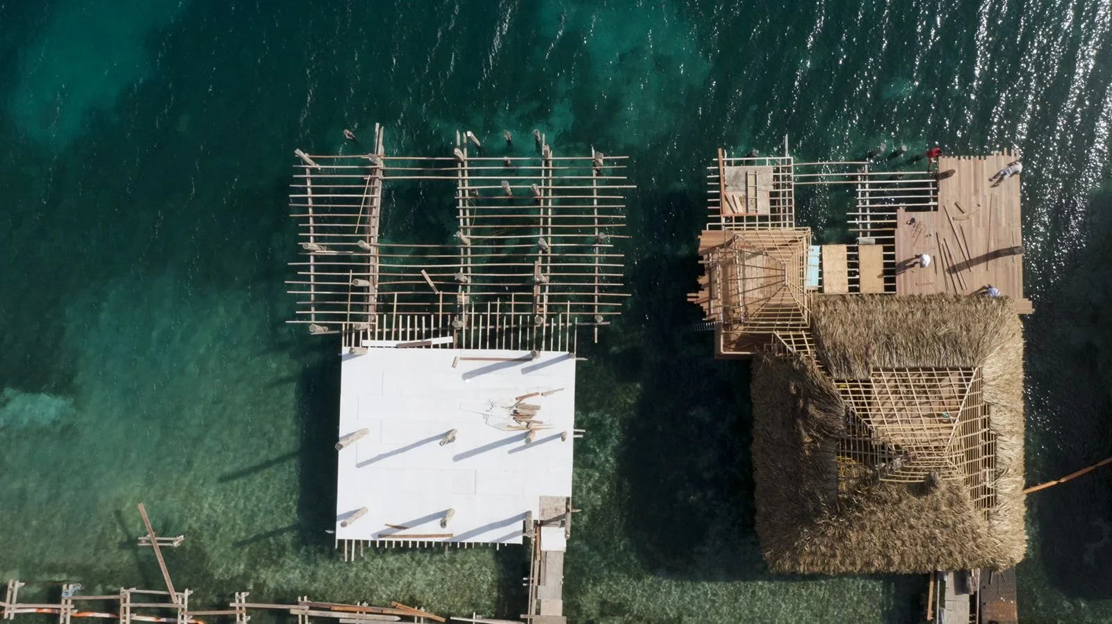Aerial view of a construction site on water showing wooden structures and thatched roofs being built.
