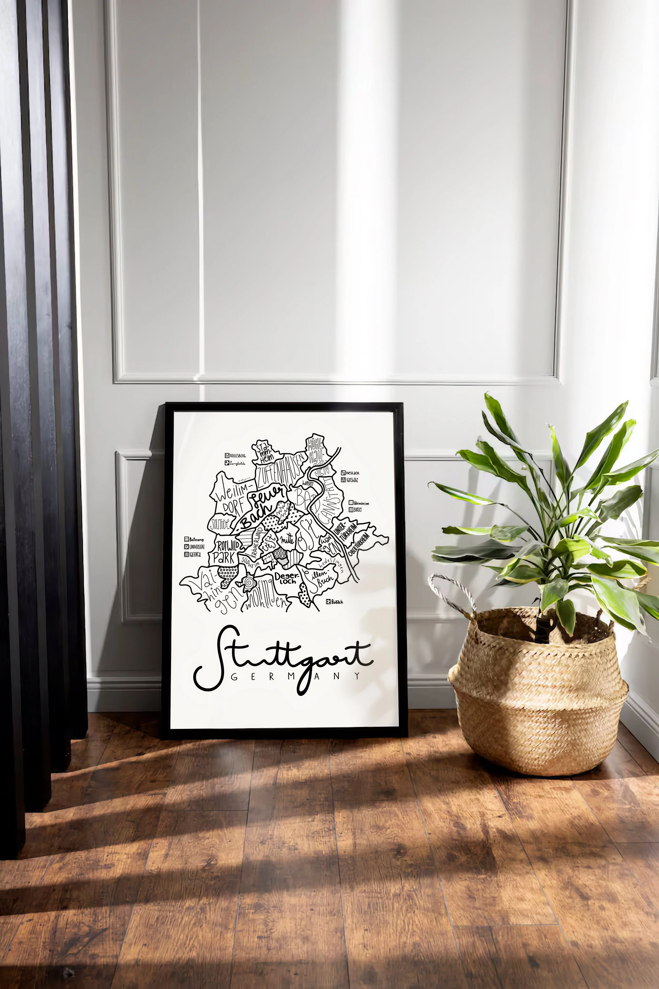 Black wood framed illustrated map of Stuttgart leaning against a wall in a modern apartment interior.