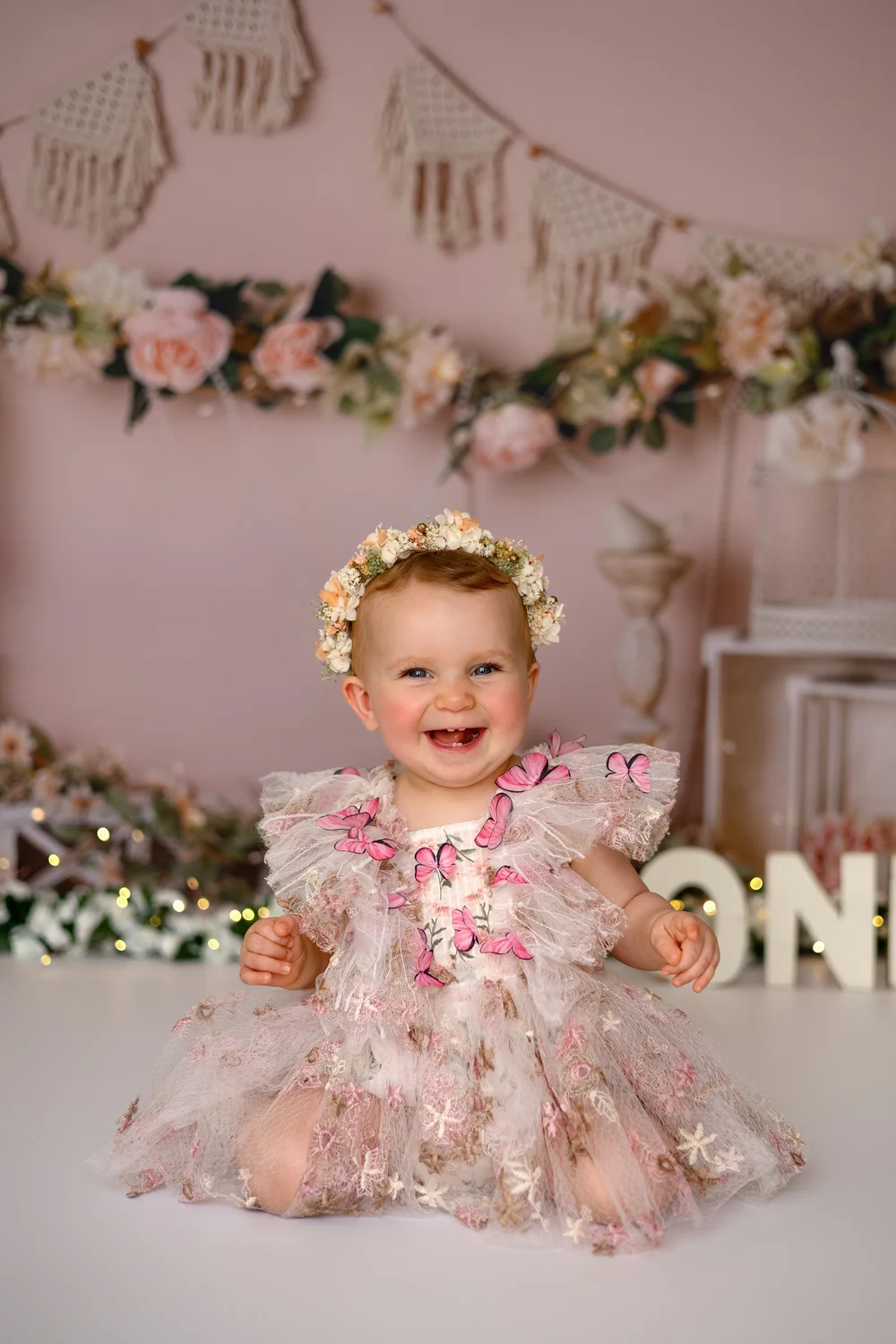 Bébé en robe fleurie et couronne de fleurs devant un décor de premier anniversaire.