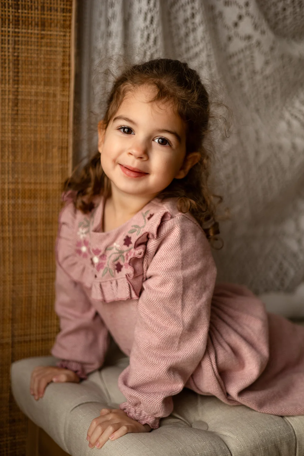 Petite fille souriante en tenue rose, assise sur une banquette capitonnée.
