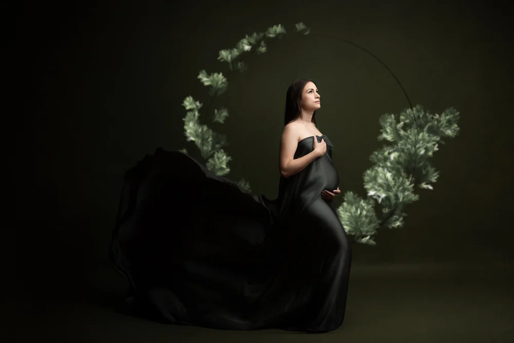 Femme enceinte en robe noire sous une arche de feuillage, sur fond sombre.