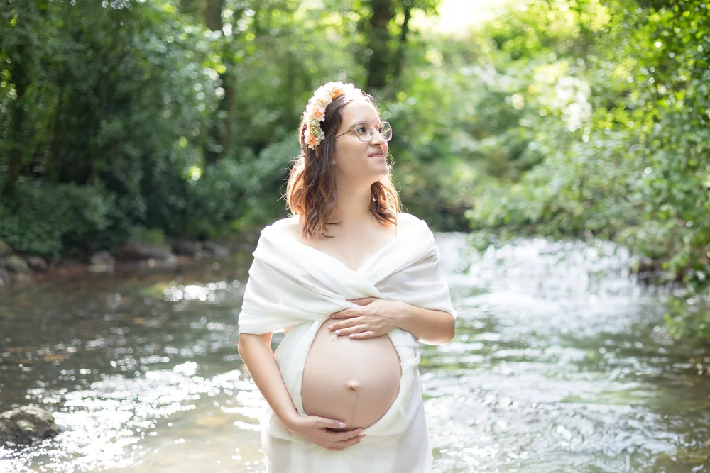 Femme enceinte en robe blanche dans une rivière, couronne de fleurs.