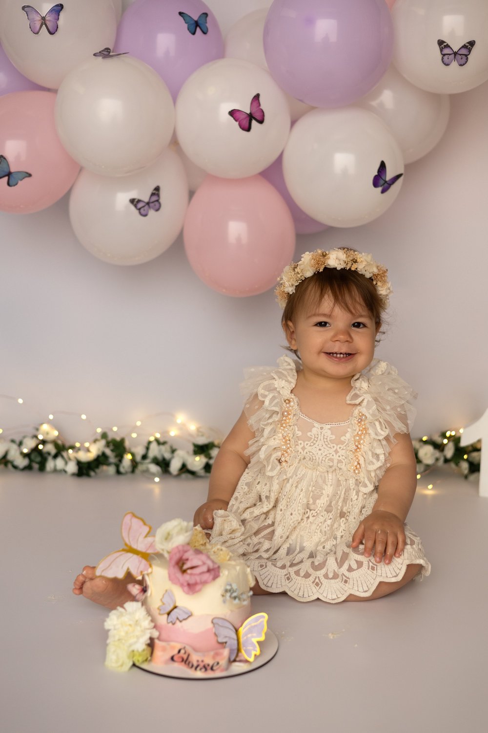 Bébé en robe de dentelle devant une arche de ballons roses et des papillons.