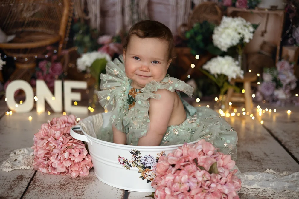 Bébé en robe verte dans une bassine fleurie, lettres « ONE » lumineuses.