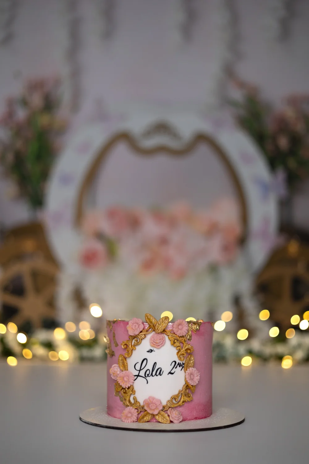 Gâteau d’anniversaire rose et doré devant une arche fleurie.