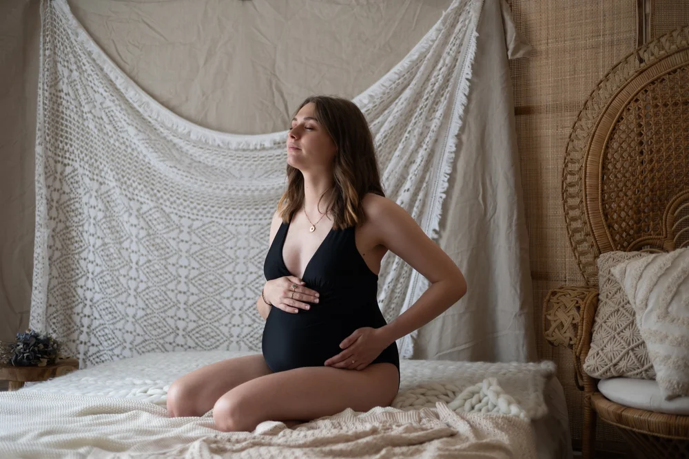 Femme enceinte en body noir assise sur un lit, en studio.