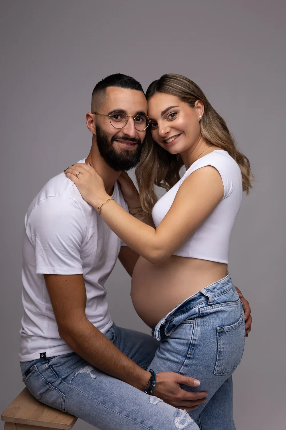 Couple assis en studio, la femme enceinte en jean et haut blanc.