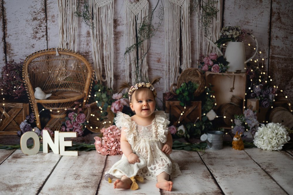Bébé en robe de dentelle devant un décor bohème, lettres « ONE ».