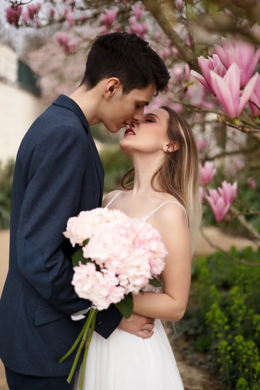 Couple s’embrassant sous un magnolia, elle tenant un bouquet d’hortensias.