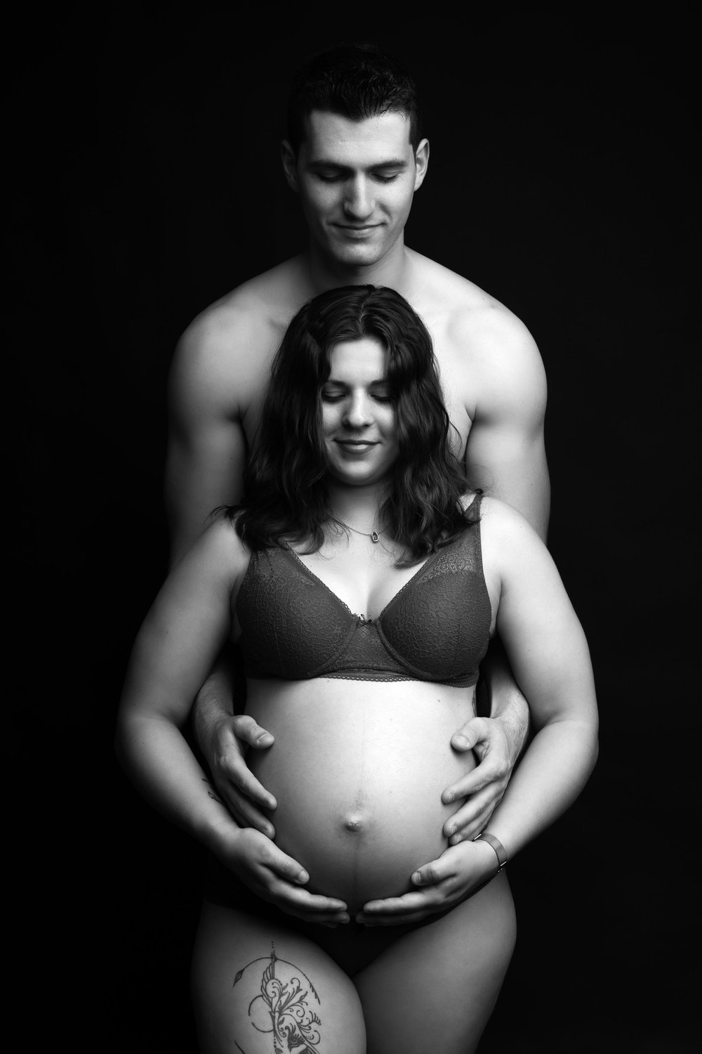 Couple en noir et blanc, mains sur le ventre, sur fond sombre.