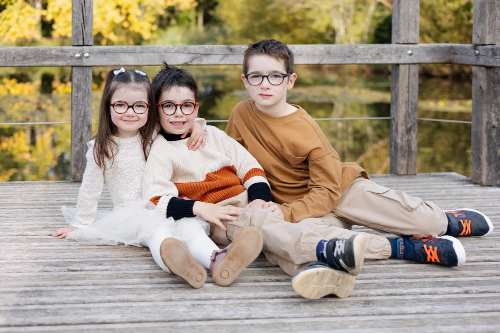 Trois enfants à lunettes assis sur un ponton en bois devant un étang.