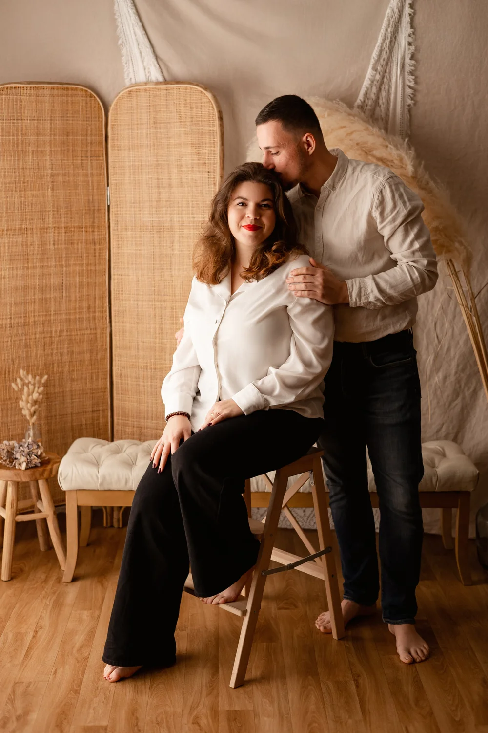 Couple dans un décor bohème, elle assise sur un tabouret, lui l’embrassant sur la tempe.
