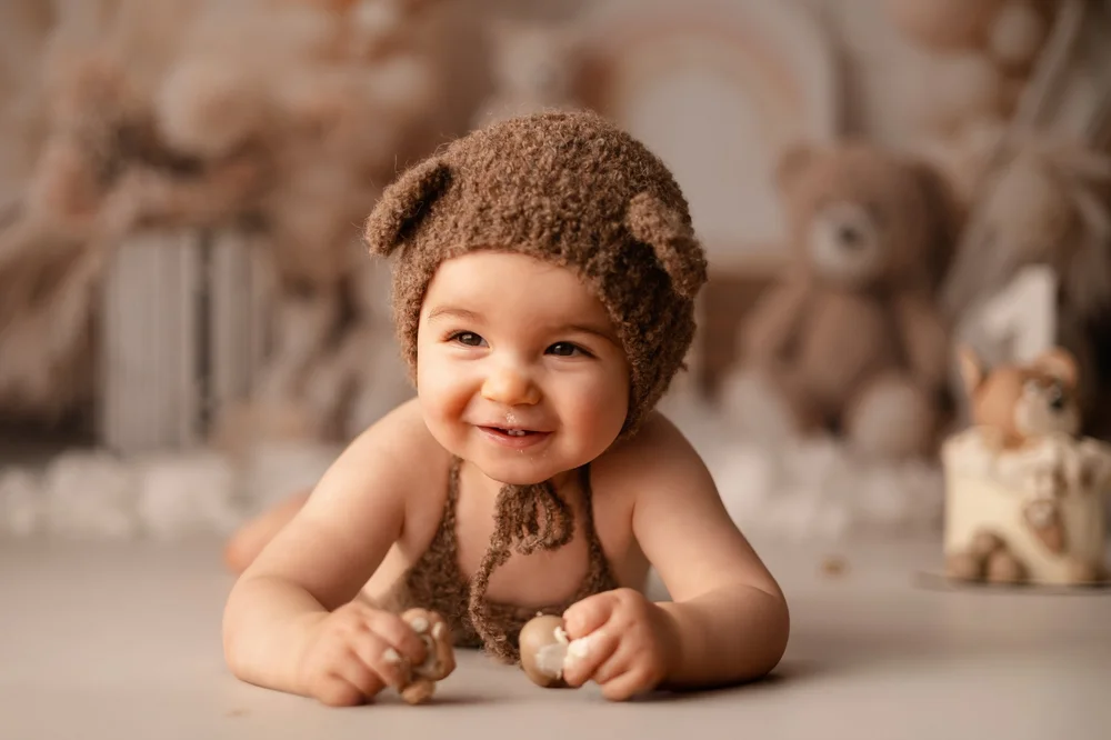 Bébé souriant coiffé d’un bonnet d’ourson, allongé devant des peluches.