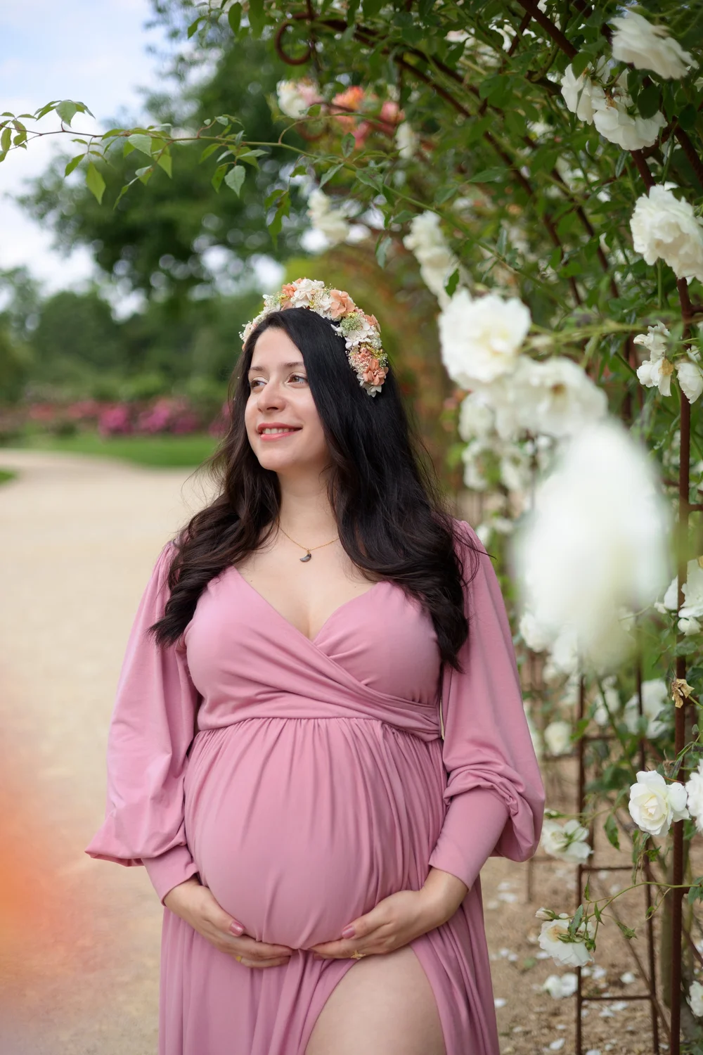 Femme enceinte en robe rose, couronne de fleurs, sous une arche de roses.