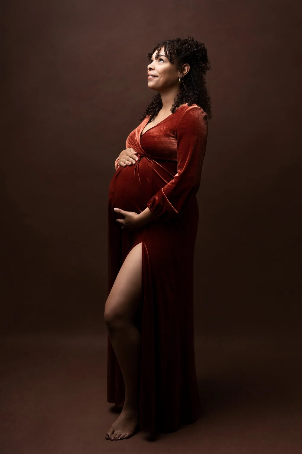 Femme enceinte en robe de velours rouille, de profil, sur fond brun.