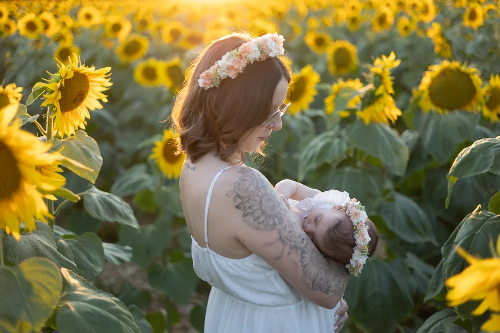 Maman couronnée de fleurs tenant son bébé dans un champ de tournesols.