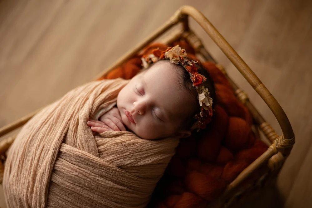 Nouveau-né endormi emmailloté de beige dans un lit en rotin, avec une couronne de fleurs.