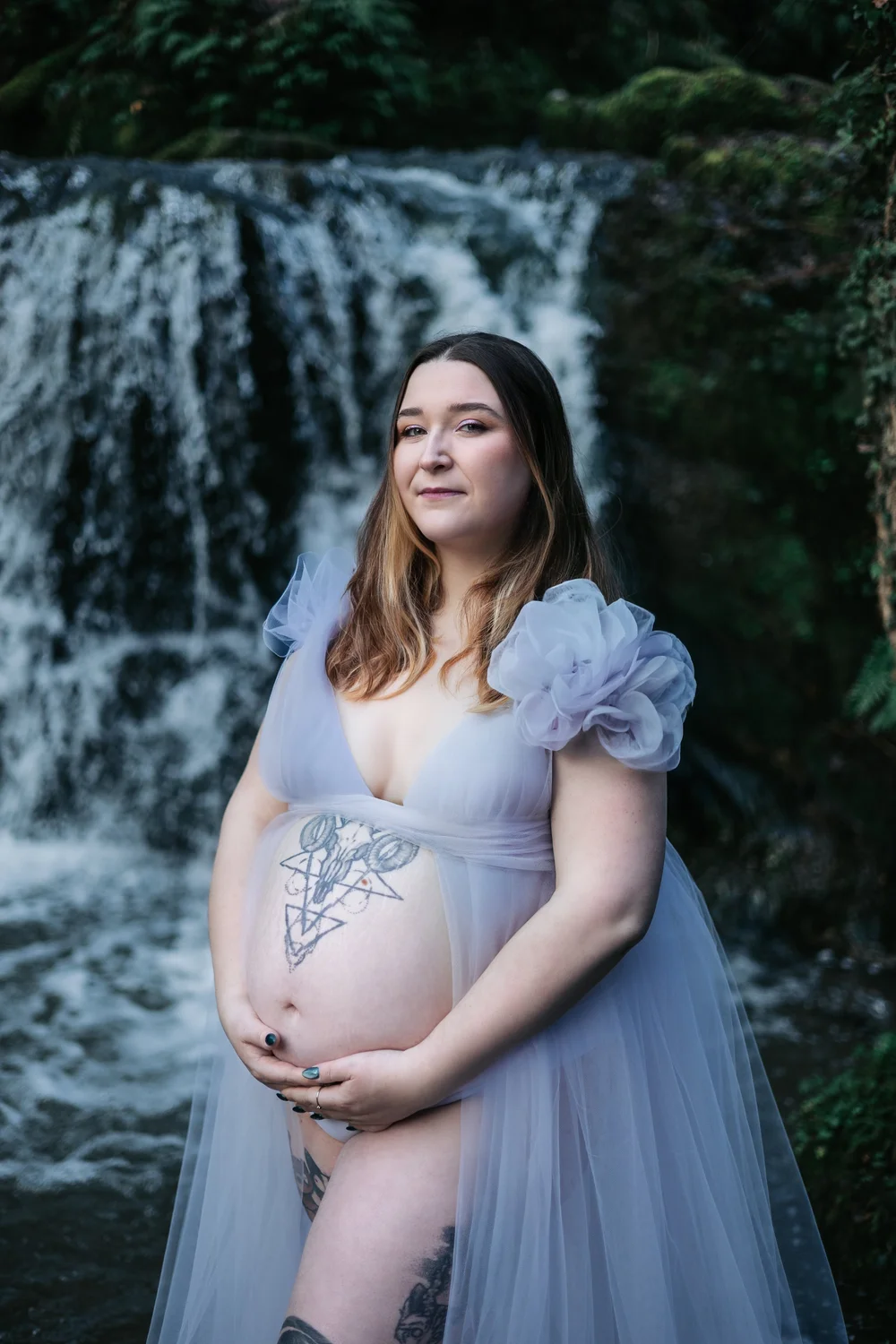 Femme enceinte en robe bleu pâle devant une cascade.