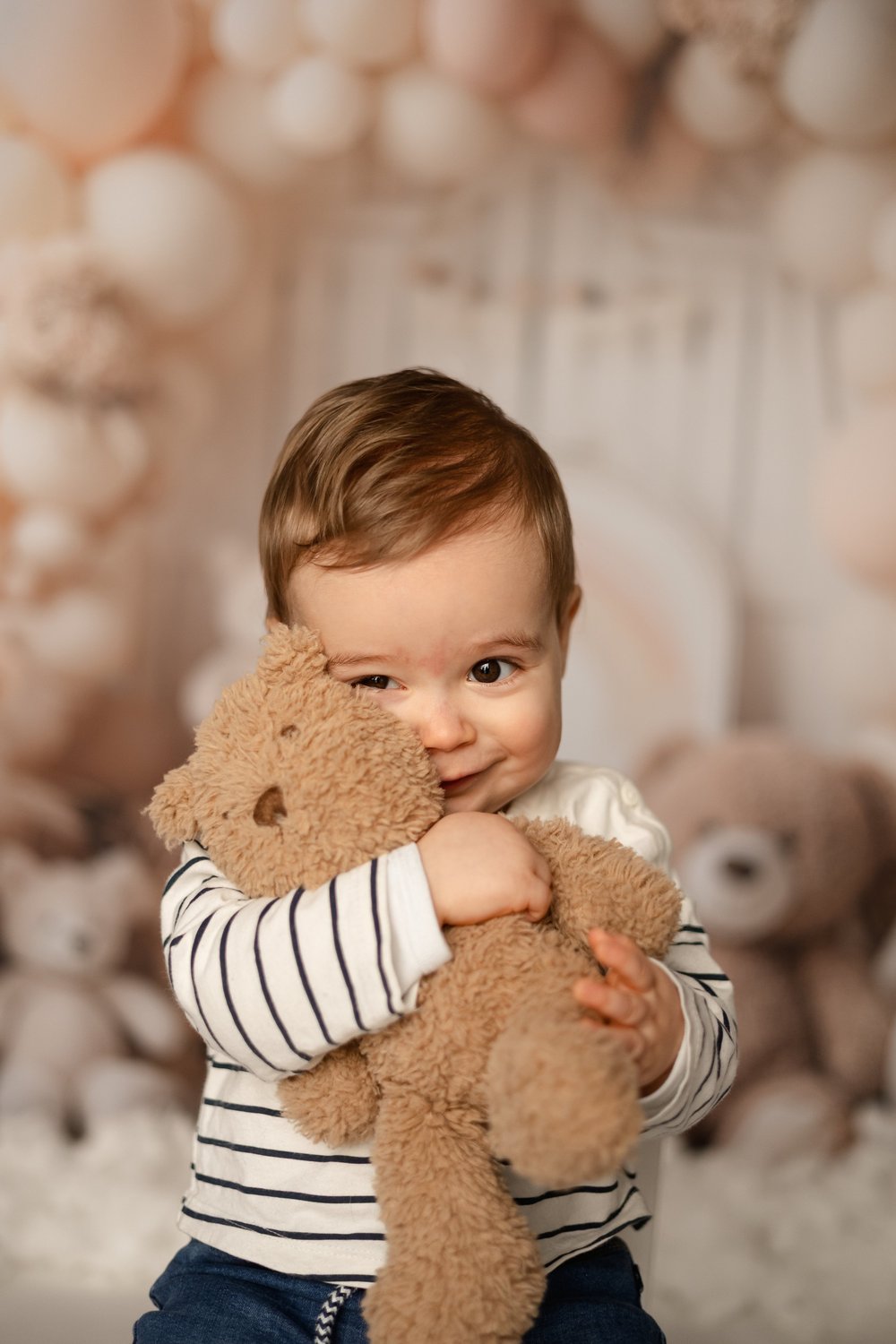 Petit garçon souriant serrant un ours en peluche, devant des ballons beiges.
