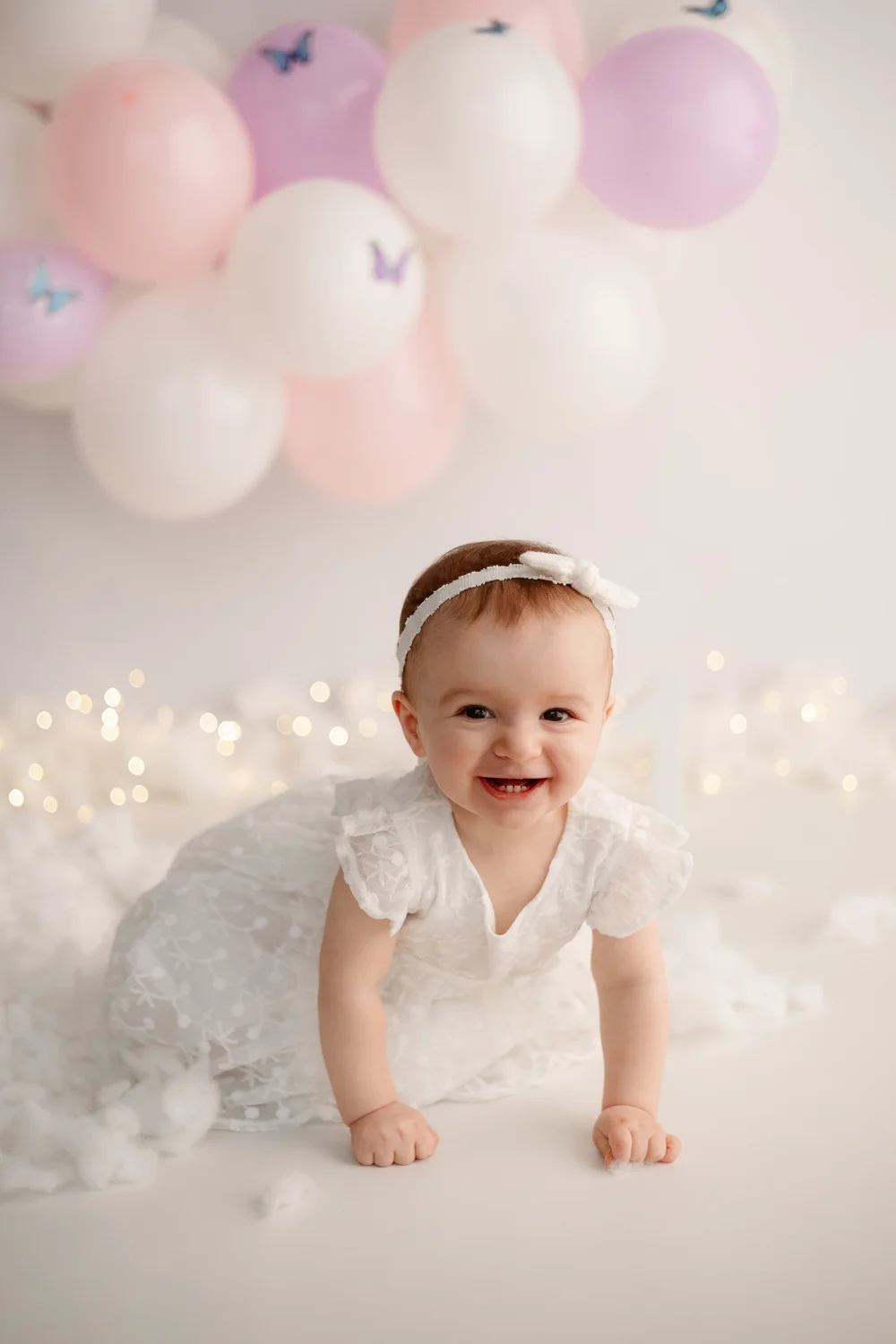 Bébé à quatre pattes en robe blanche devant des ballons roses et blancs.