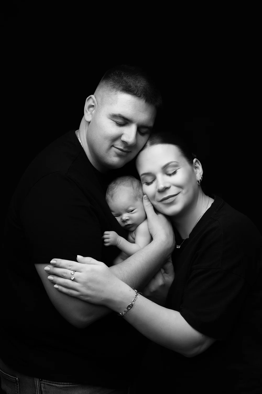 Photo en noir et blanc : parents serrant leur nouveau-né contre eux, sur fond noir.