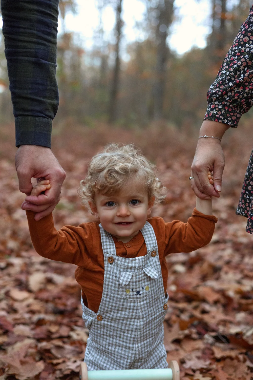 Bébé marchant dans les feuilles d’automne, tenu par les mains de ses parents.