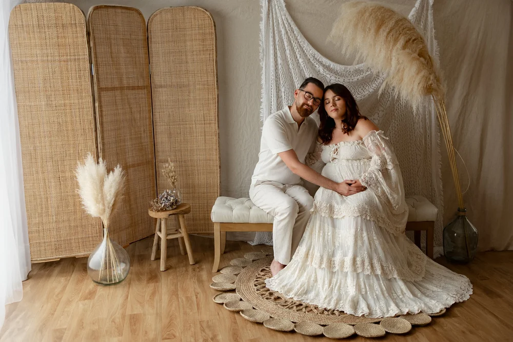 Couple assis en studio devant un paravent en rotin, la femme enceinte en robe blanche.