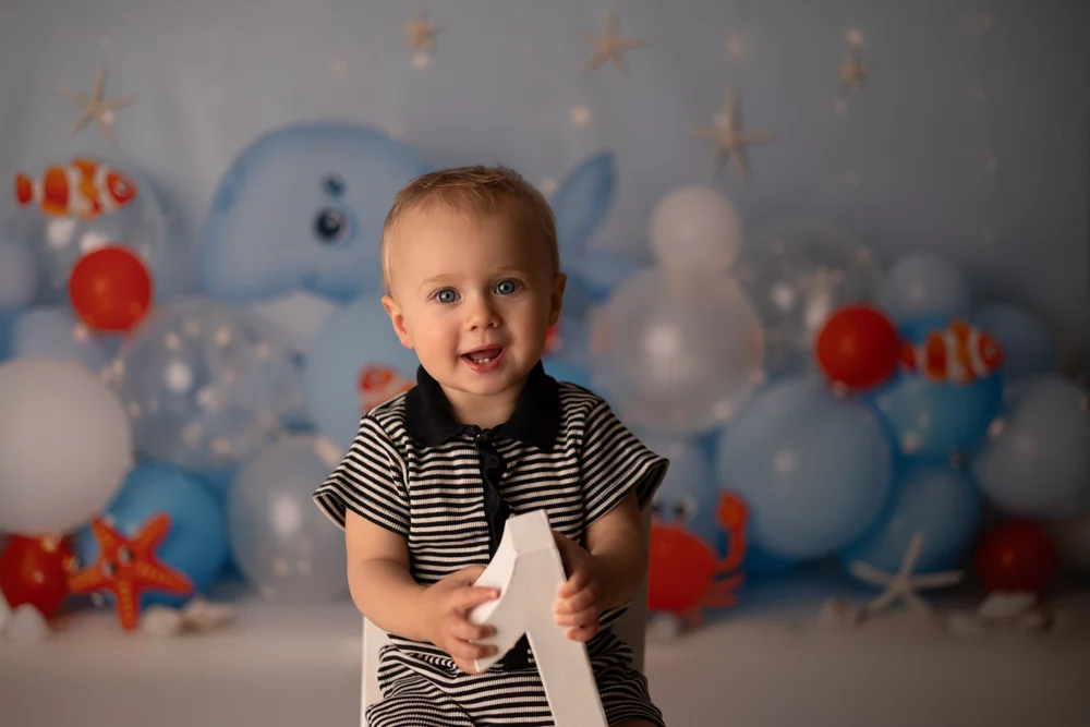 Petit garçon souriant tenant un chiffre 1, devant un décor marin bleu.