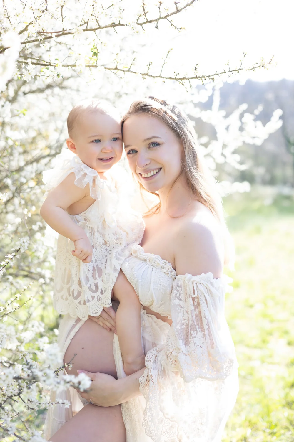 Future maman portant son bébé, en robe de dentelle, sous un arbre en fleurs.