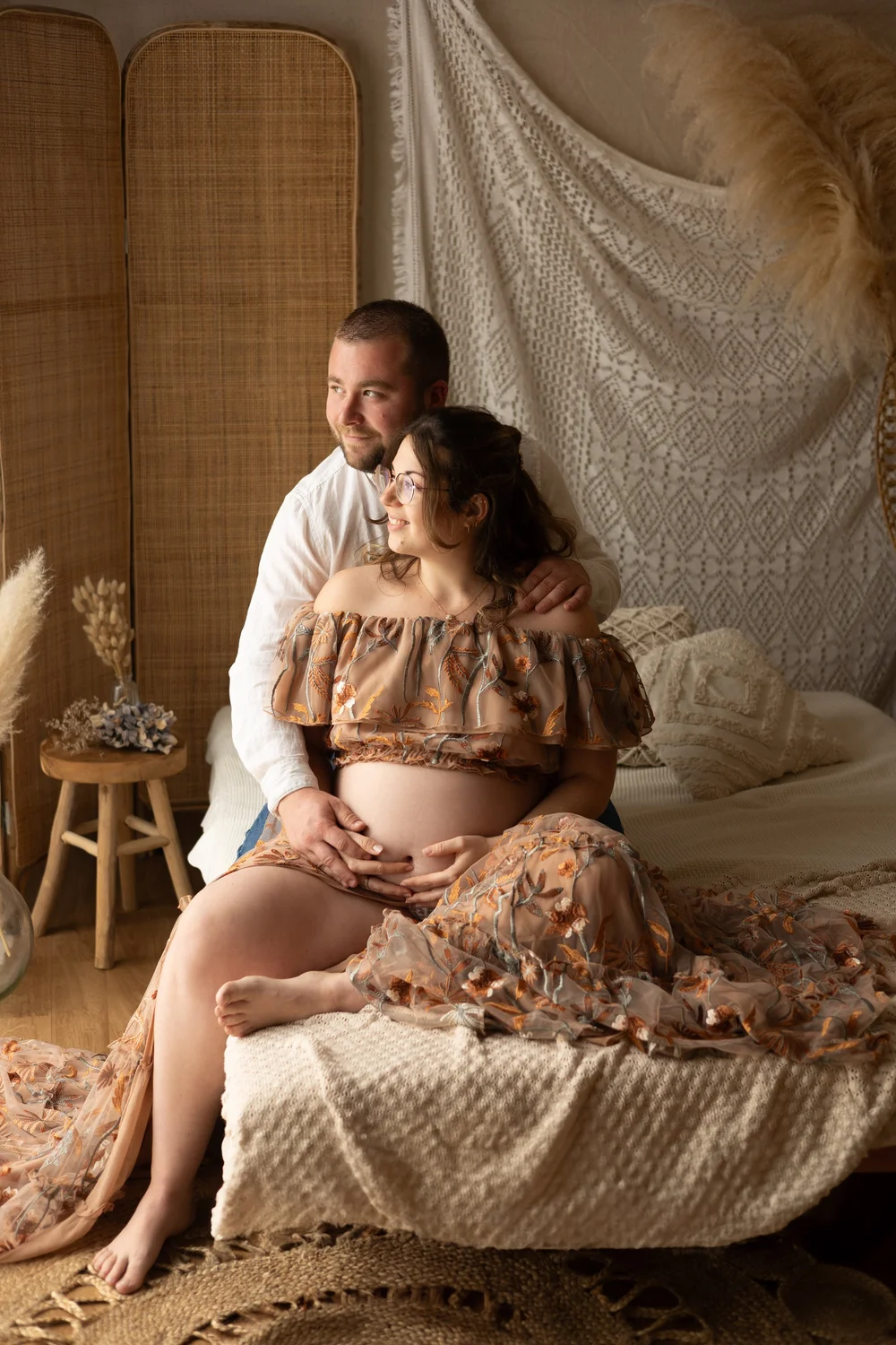 Couple assis sur un lit en studio, la femme enceinte en robe fleurie.