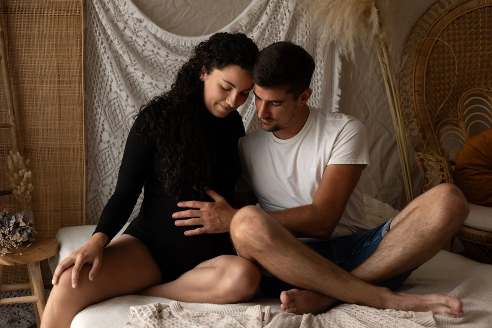 Couple assis sur un lit en studio, la femme enceinte en robe noire.