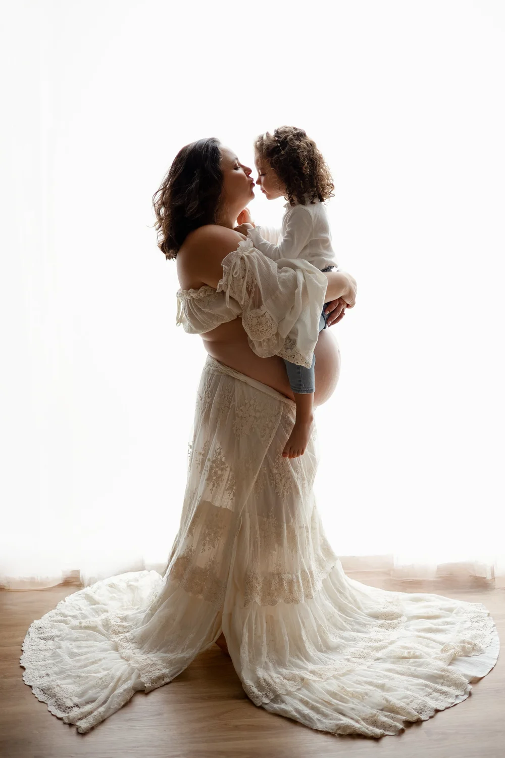Future maman portant sa fille dans les bras, en contre-jour sur fond blanc.