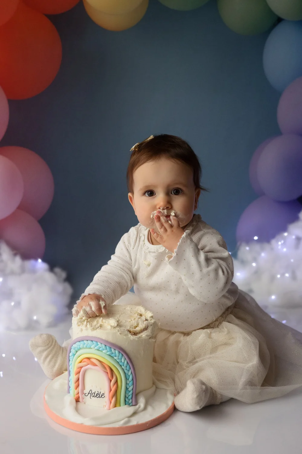Bébé en robe blanche goûtant un gâteau arc-en-ciel, ballons pastel.