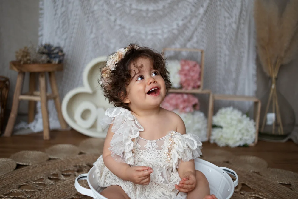 Bébé en robe blanche devant un cœur « ONE » et des fleurs.