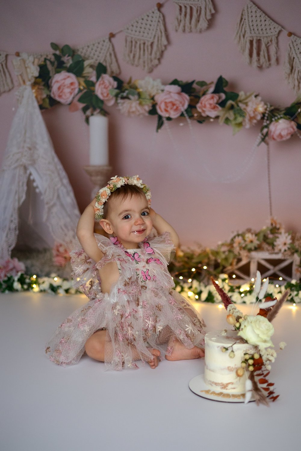 Bébé en robe fleurie, couronne de fleurs, devant un décor rose pour son anniversaire.