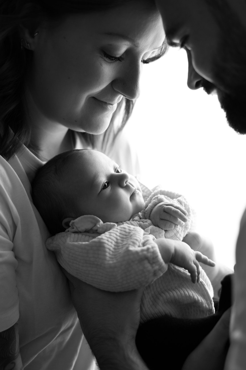 Parents penchés sur leur nouveau-né, en noir et blanc, en contre-jour.