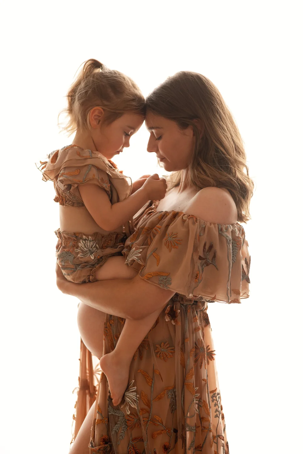 Future maman et sa petite fille front contre front, en robes fleuries, sur fond blanc.