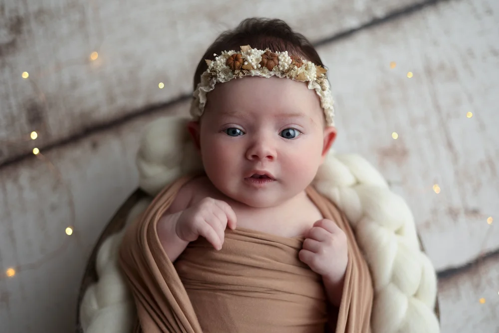 Nouveau-né les yeux ouverts, bandeau fleuri, emmailloté de beige.