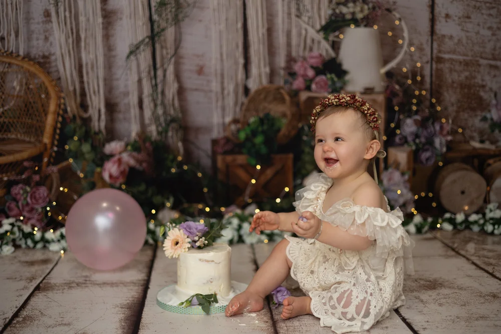 Bébé en robe de dentelle avec un ballon rose et son gâteau, décor fleuri.