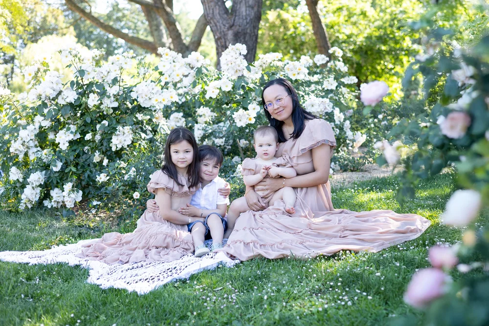 Maman et ses deux enfants assis sur un plaid, au pied de rosiers blancs.