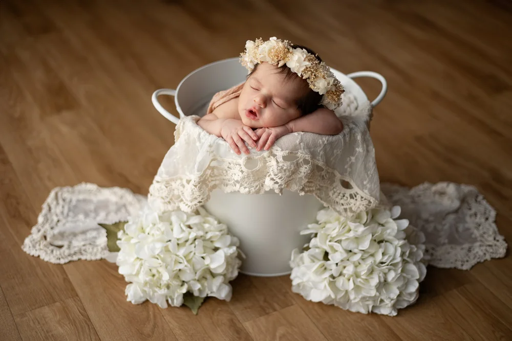 Nouveau-né avec une couronne de fleurs dans un seau blanc garni de dentelle.