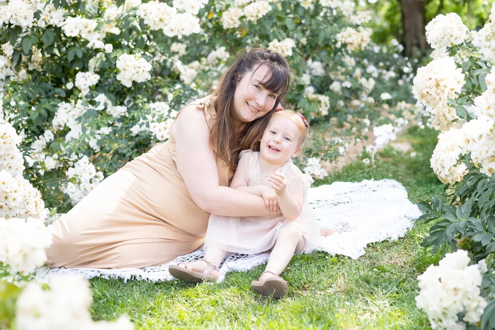 Maman assise sur un plaid avec sa fille, devant des buissons de roses blanches.