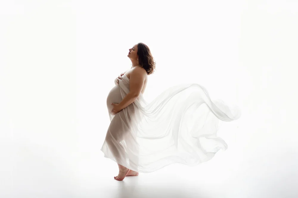 Femme enceinte en silhouette, un voile blanc flottant derrière elle, sur fond blanc.