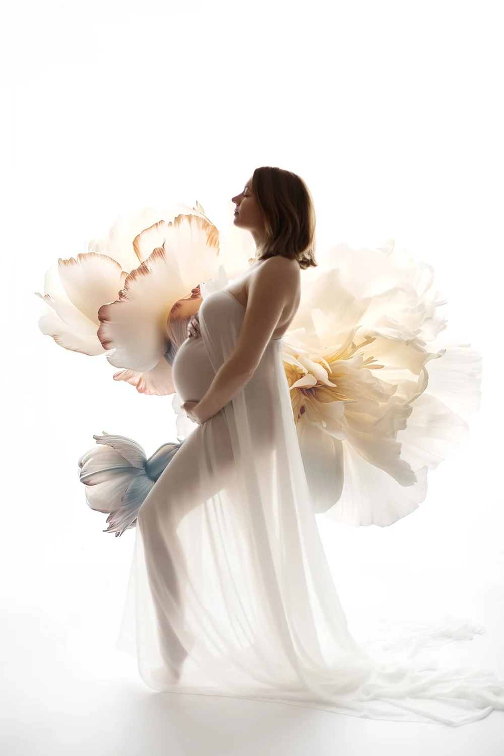 Femme enceinte en robe blanche devant de grandes fleurs en papier, sur fond clair.