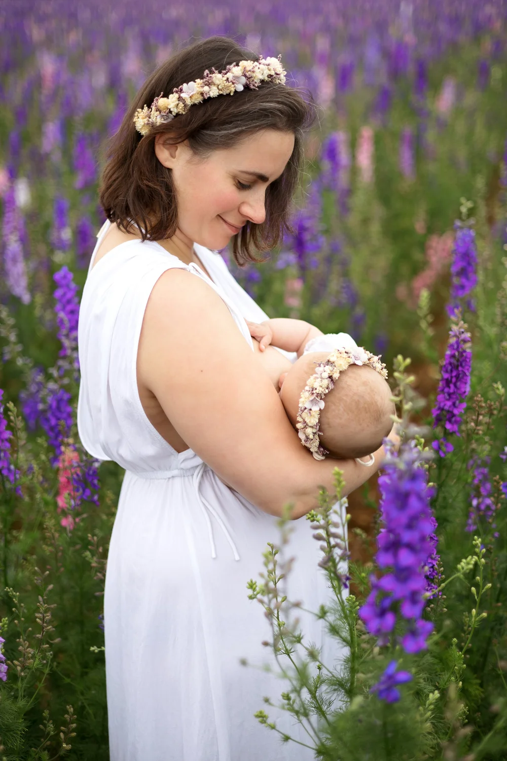 Maman en robe blanche tenant son bébé parmi des fleurs violettes.