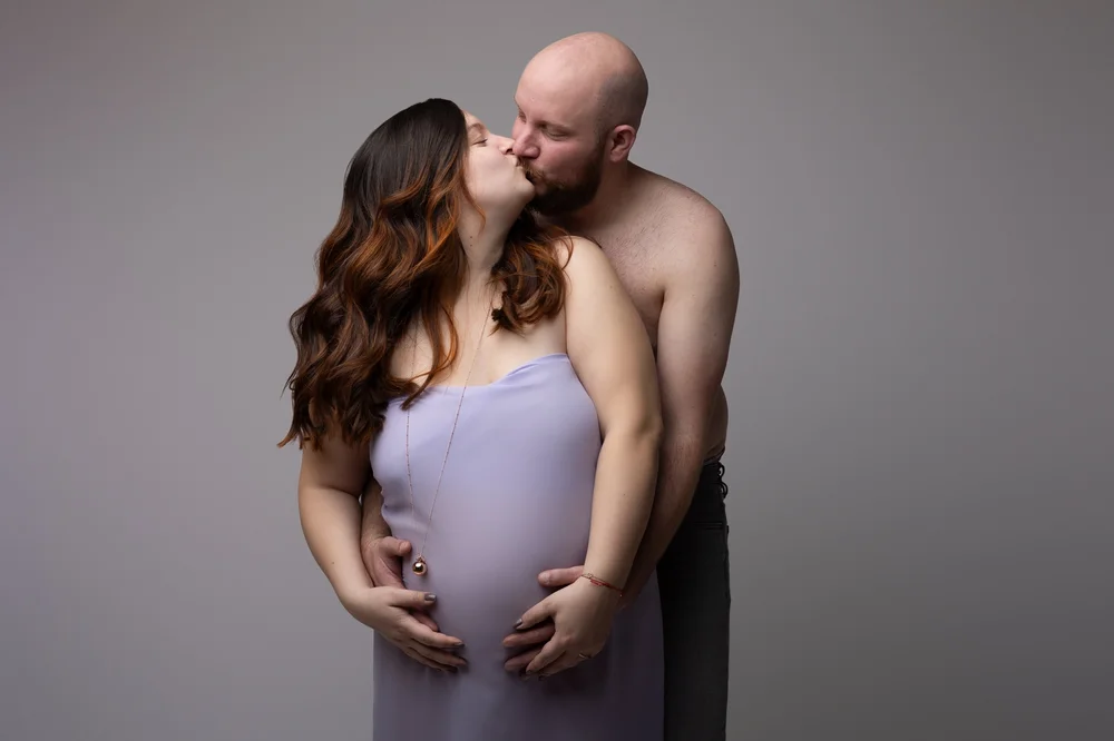 Couple s’embrassant en studio, la femme enceinte drapée de lilas.