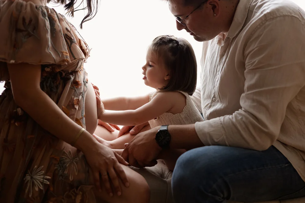 Future maman et papa avec leur petite fille, la main sur le ventre, en contre-jour.
