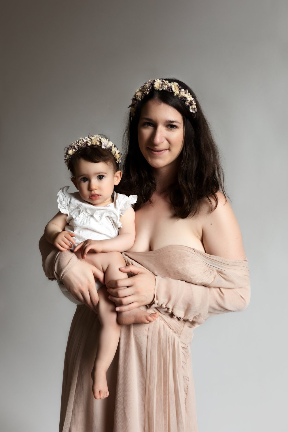 Maman en robe beige tenant son bébé, couronnes de fleurs, sur fond clair.