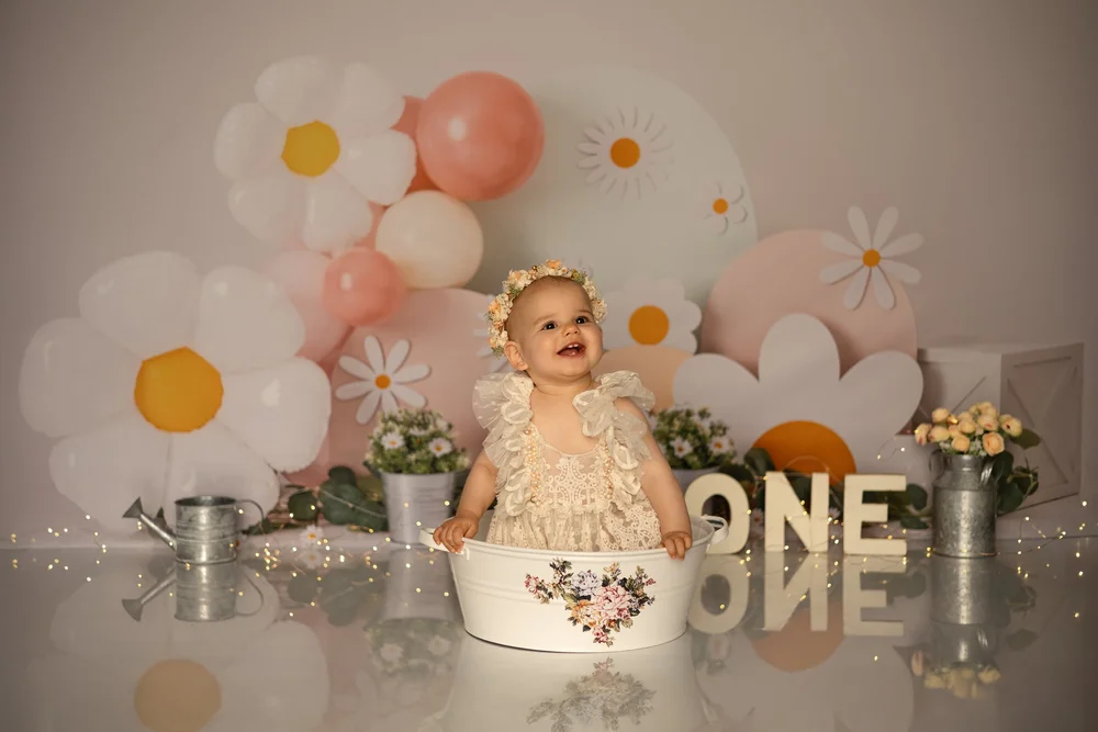 Bébé dans un bain de fleurs devant un décor de marguerites et de ballons, lettres « ONE ».
