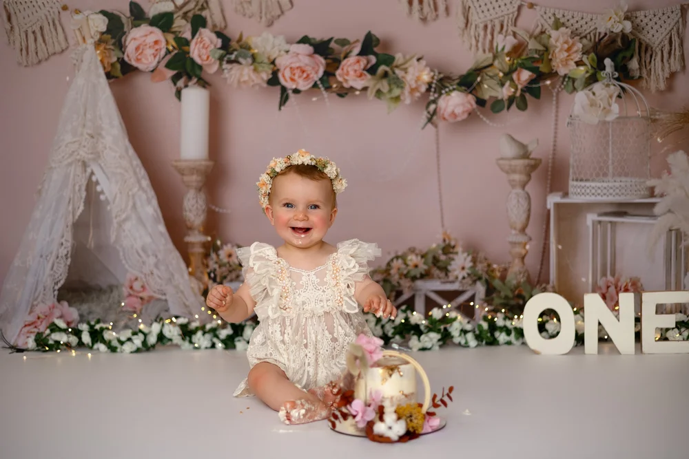 Bébé en robe de dentelle devant un décor rose fleuri, lettres « ONE ».